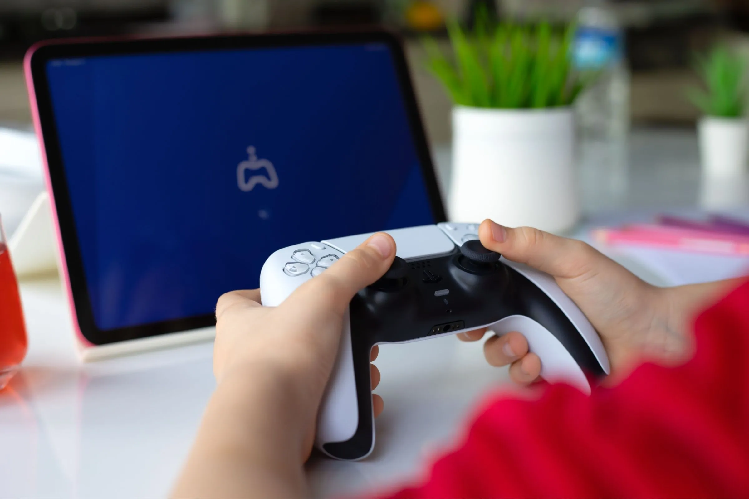 Bàn tay trẻ em cầm tay cầm DualSense PS5 và iPad Air, minh họa khả năng chơi game từ xa qua Remote Play.