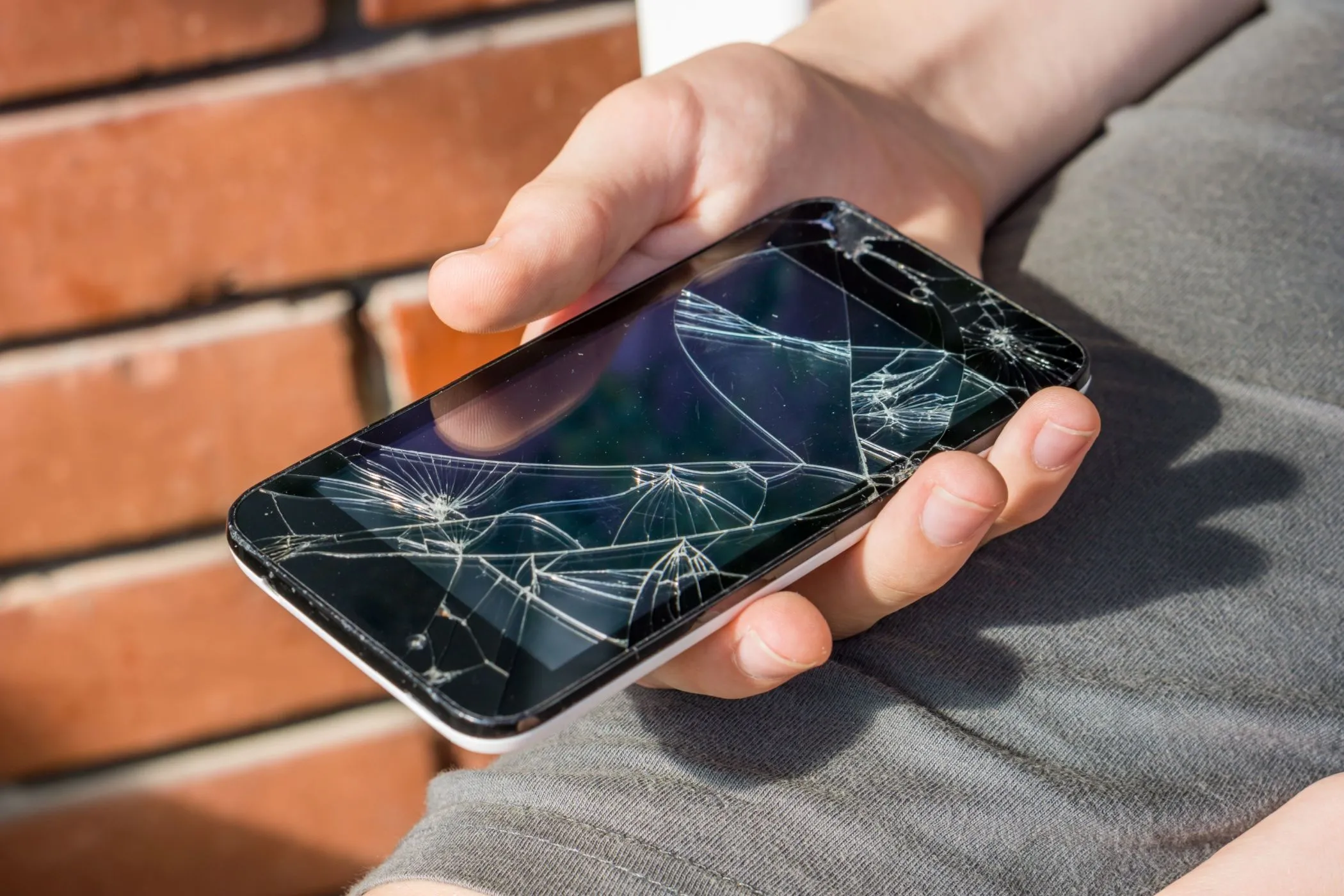 Cận cảnh bàn tay một cậu bé cầm chiếc điện thoại smartphone bị vỡ màn hình nhưng vẫn hoạt động, minh họa sự mong manh của thiết bị di động hiện đại và nhu cầu về độ bền.
